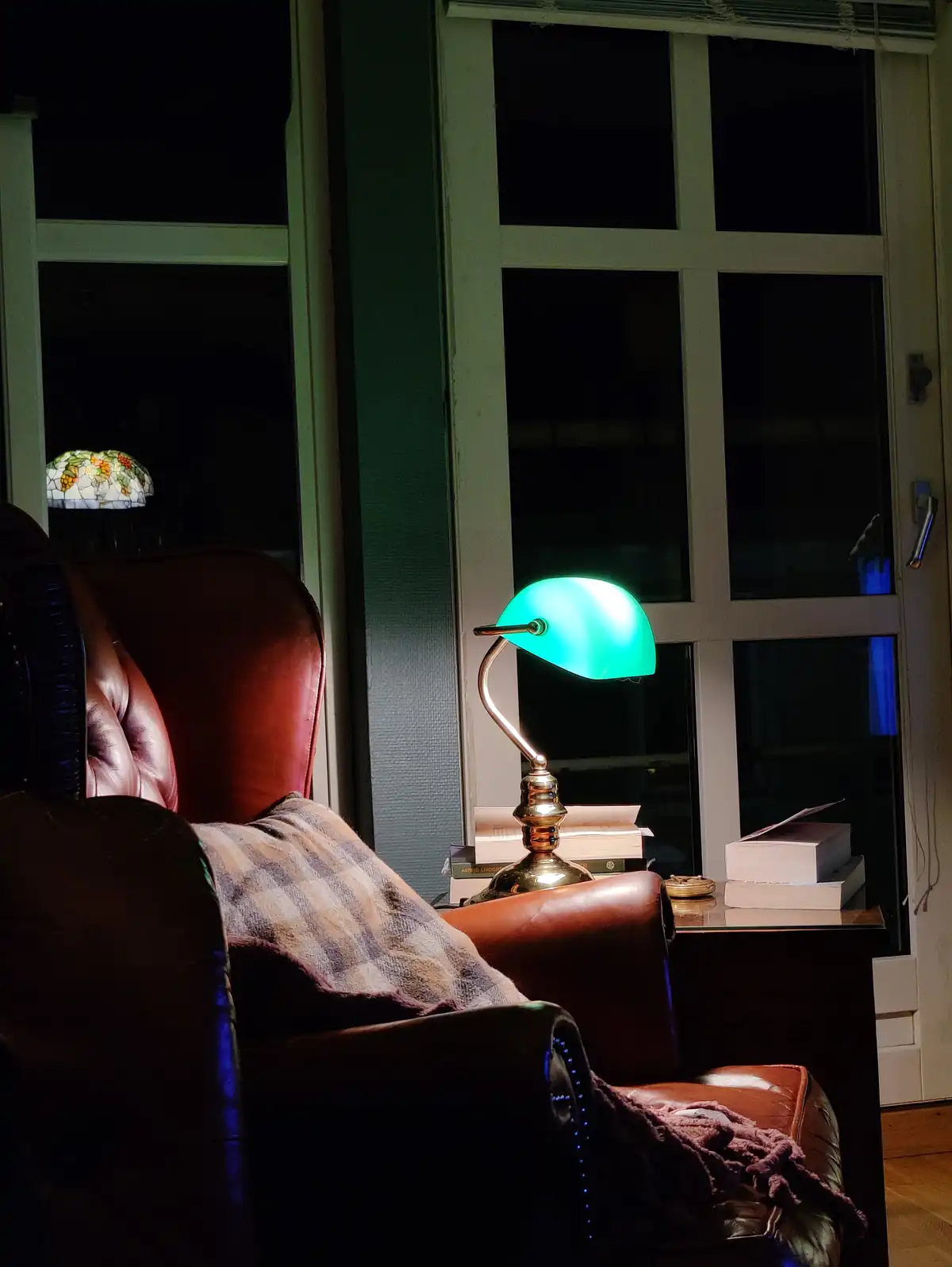 A cozy, dimly lit room at night features a brown leather armchair with a plaid blanket draped over it. A glowing green desk…