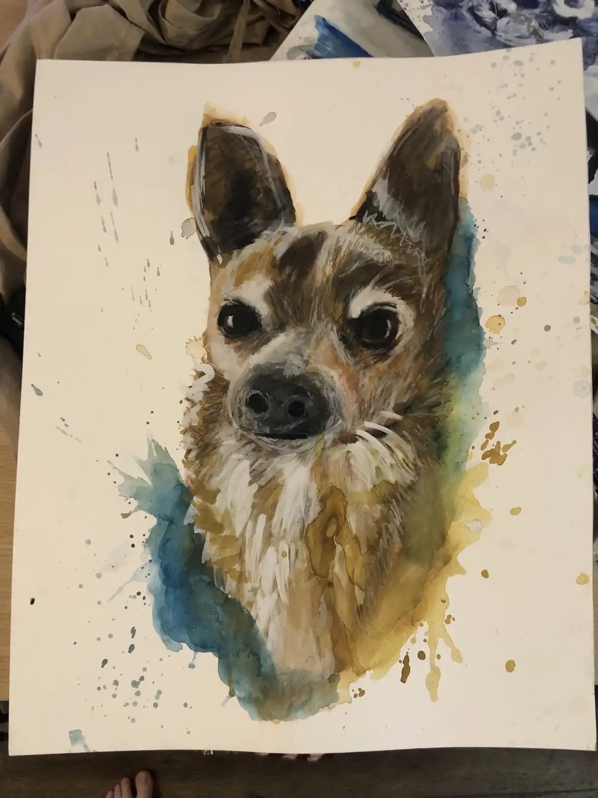 A watercolor painting of a small dog's face with brown, white, and black fur, surrounded by blue and gold paint splatters on…