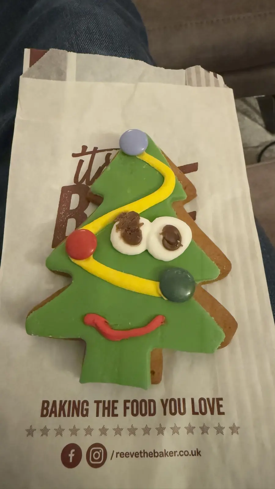 A gingerbread cookie shaped like a Christmas tree with green icing, a yellow candy garland, and a red smile. It has white…