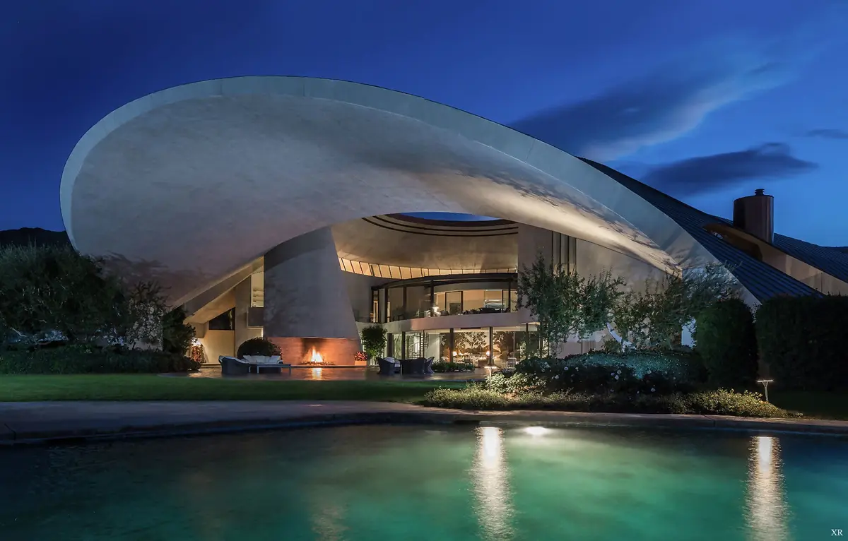 a stunning nighttime photograph of a futuristic, curved concrete house with a large pool in the foreground, illuminated by…