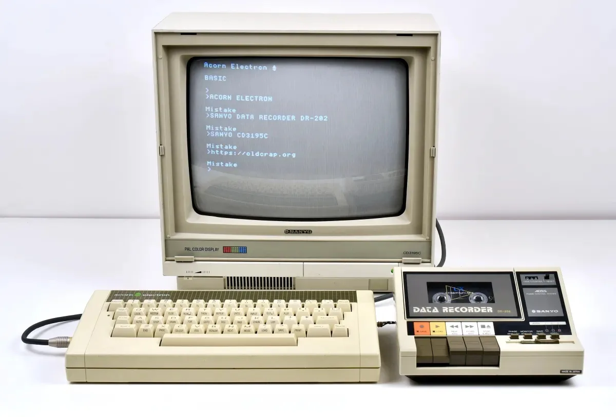 An Acorn Electron computer with a Sanyo data recorder attached, its screen displays a BASIC prompt followed by three…