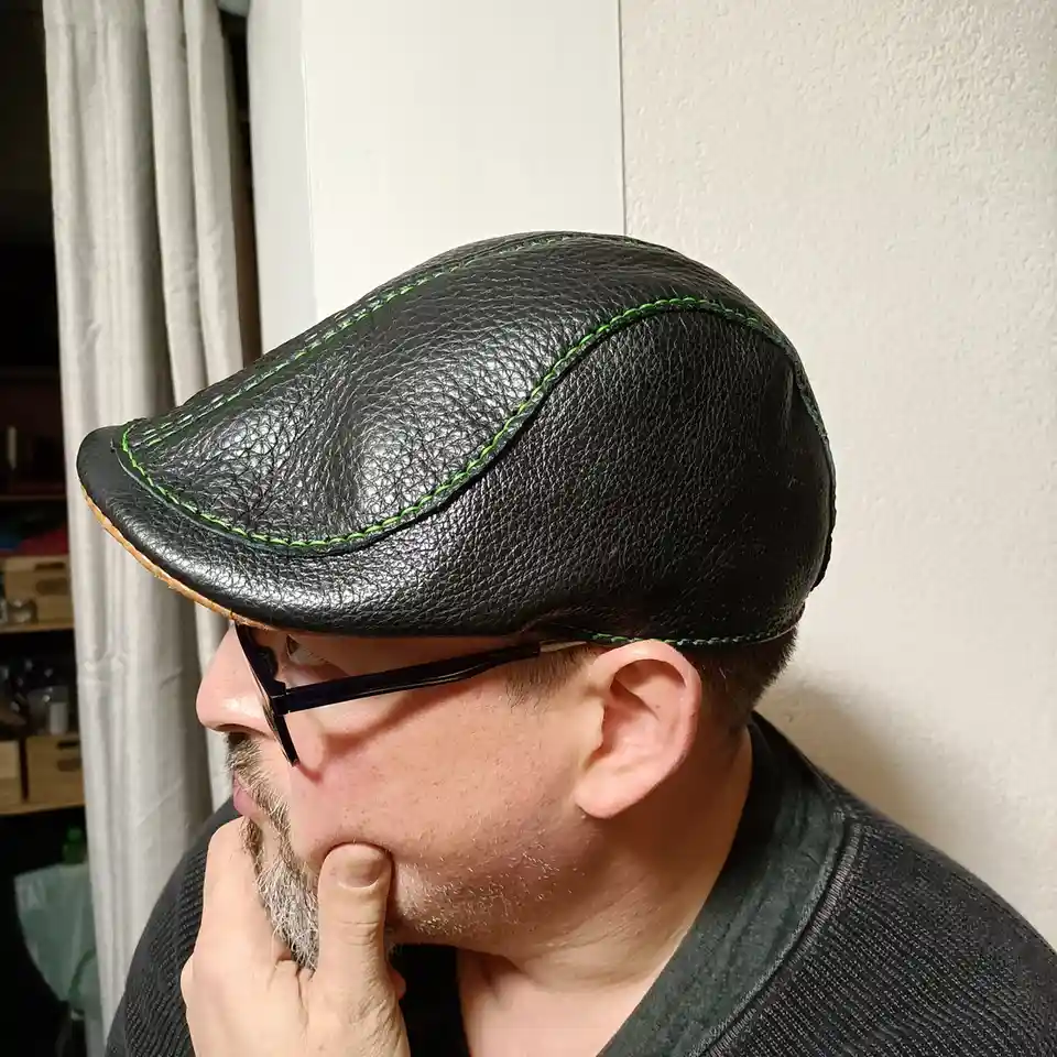 A man in profile wearing a black leather flat cap with bright green stitching, looking thoughtful with his finger on his…