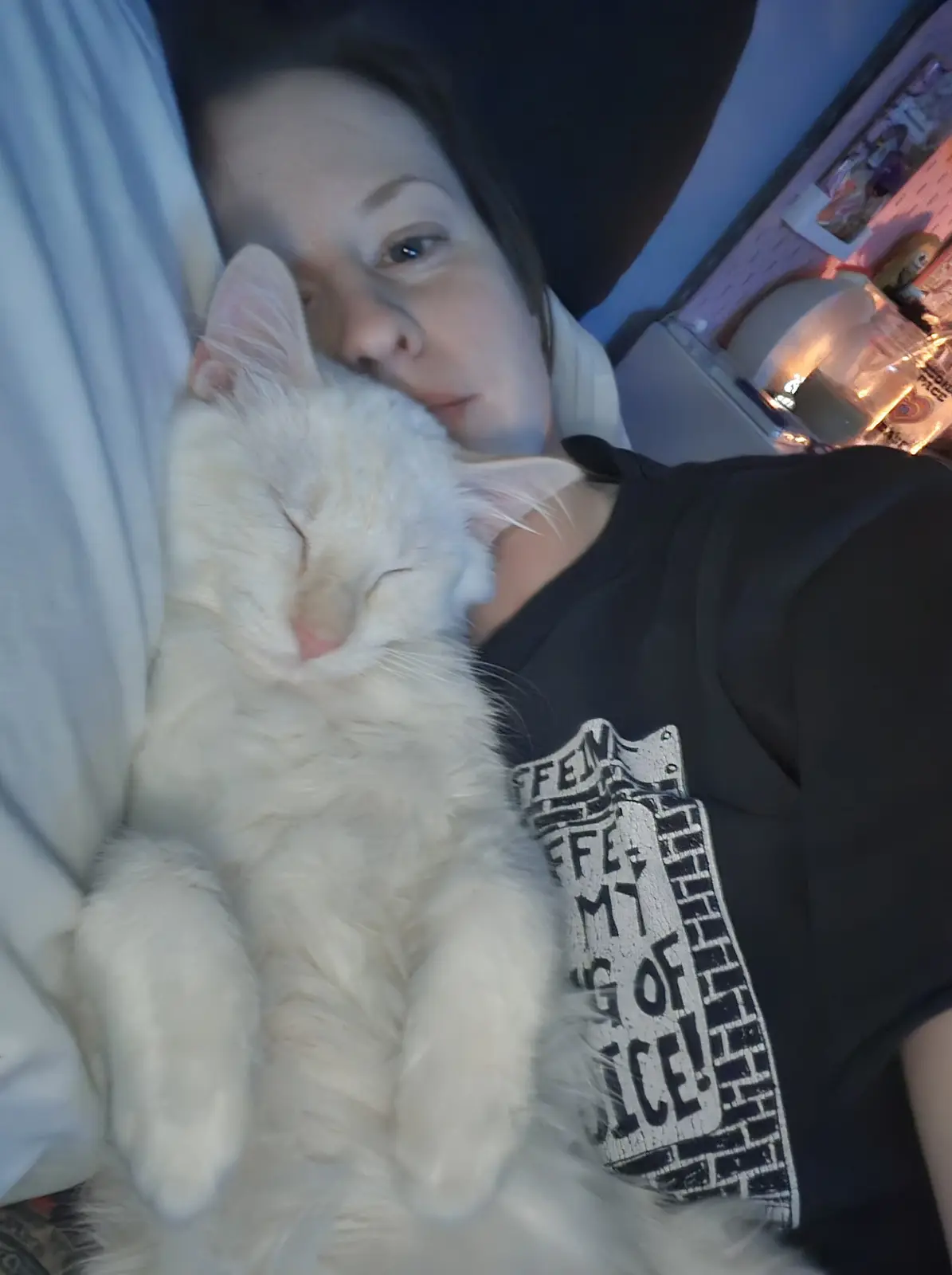 A person is lying in bed, cuddling a fluffy white cat that is sleeping soundly with its eyes closed. The person is wearing a…