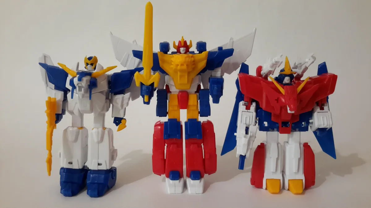 Three plastic robot toys stand side-by-side against a plain white background. The central robot is larger, with a yellow…