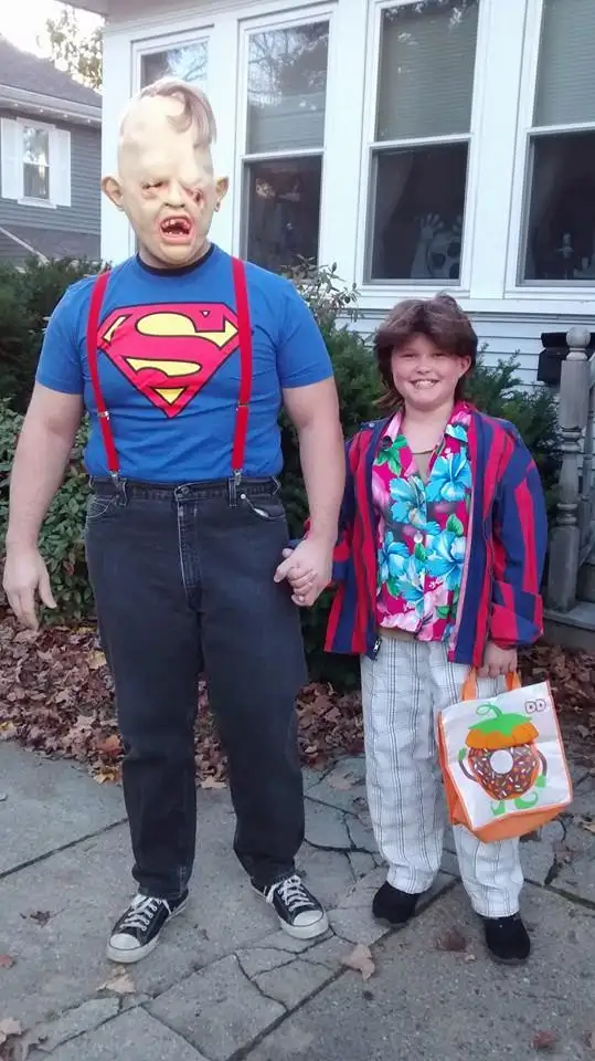 Two people stand outside a house holding hands. The person on the left wears a blue Superman t-shirt with red suspenders…