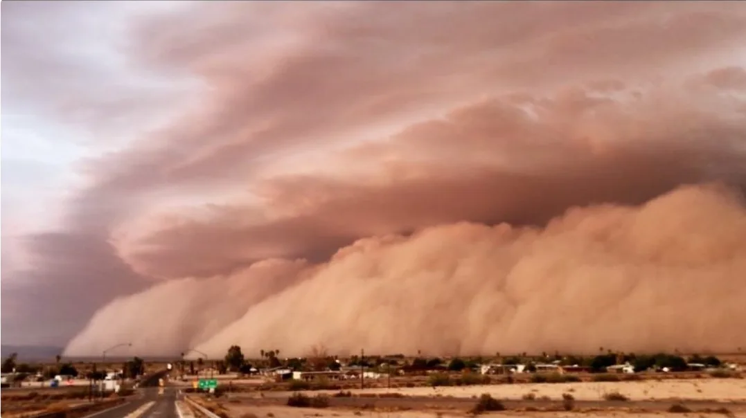 A wide desert landscape shows a massive, ominous wall of dust and sand moving across the horizon, obscuring the sky and…