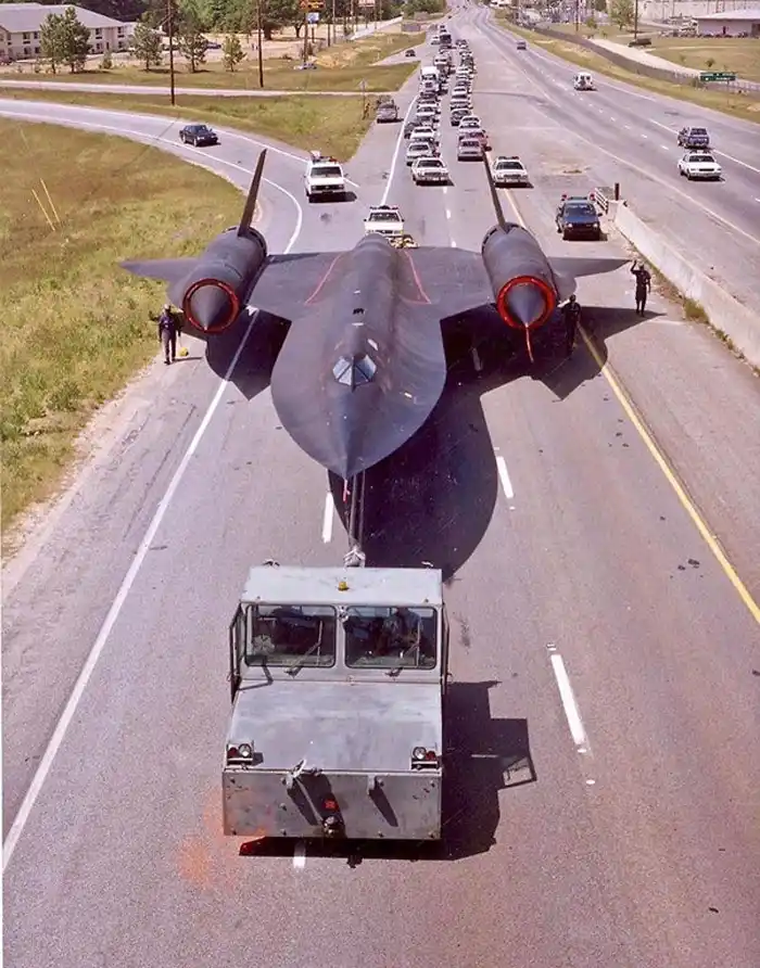 A black, stealthy SR-71 Blackbird reconnaissance aircraft is being transported down a multi-lane highway on a flatbed truck…