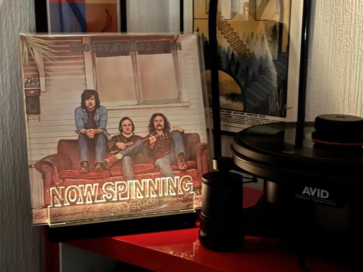 A vinyl record album cover of Crosby, Stills & Nash is displayed upright on a red shelf next to a black AVID turntable. The…