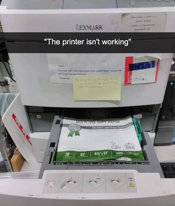 A Lexmark printer has its paper tray open, revealing a ream of copy paper. A handwritten note taped to the printer says…