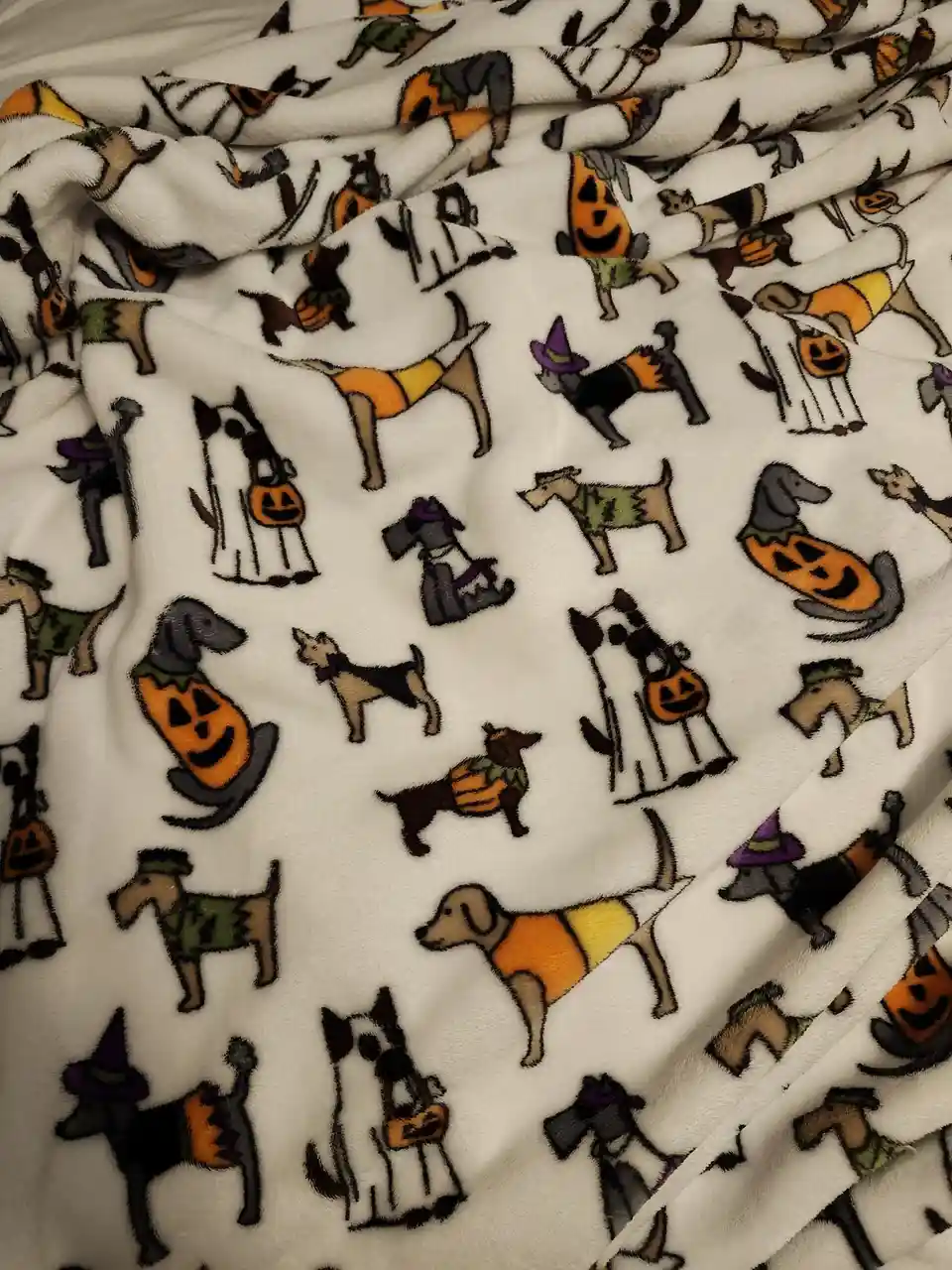 A close-up of a white, fluffy blanket with a repeating pattern of cartoon dogs dressed in various Halloween costumes…