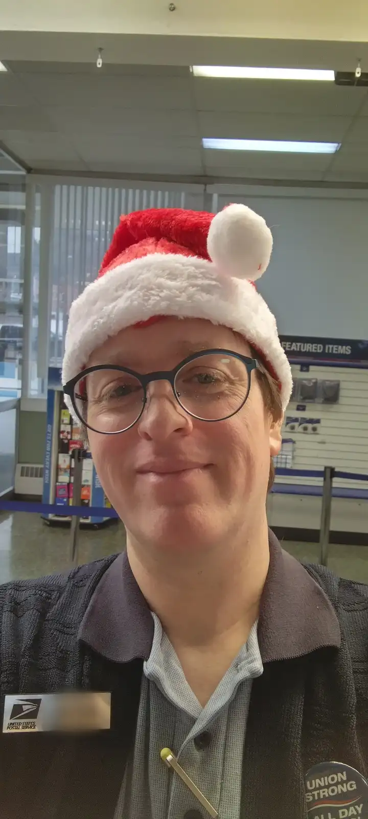 A smiling postal worker wearing a red and white Santa hat and glasses, standing inside a United States Postal Service…
