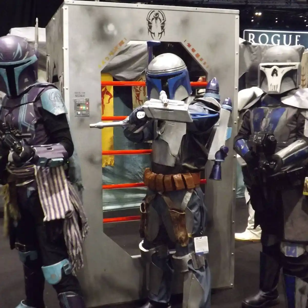 Three people in Mandalorian armor cosplay stand beside a booth with a 'ROGUE' sign. The central figure, in blue and silver…