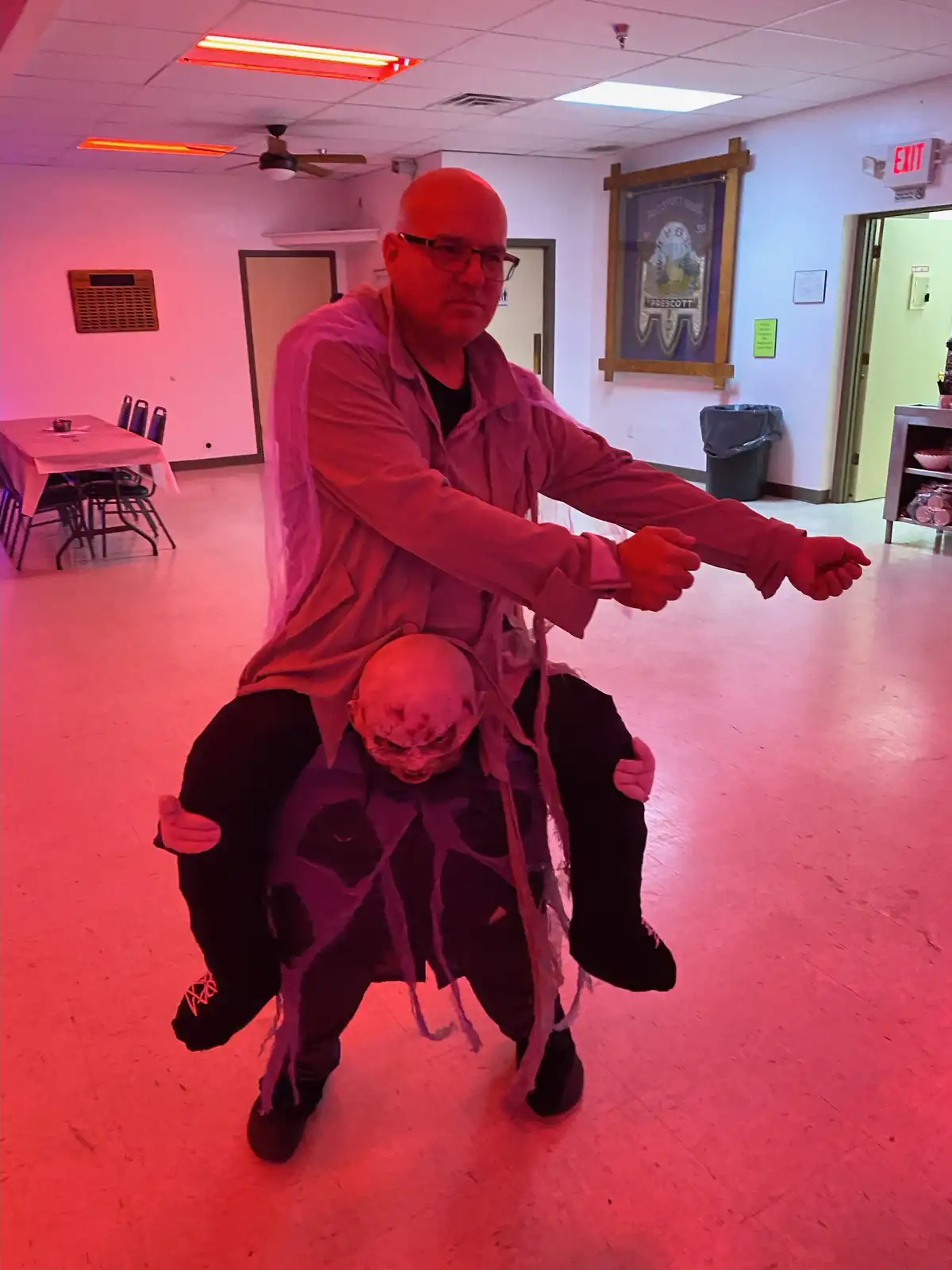A man in a red-lit room is posing with arms outstretched while sitting atop another person wearing a demonic costume with a…