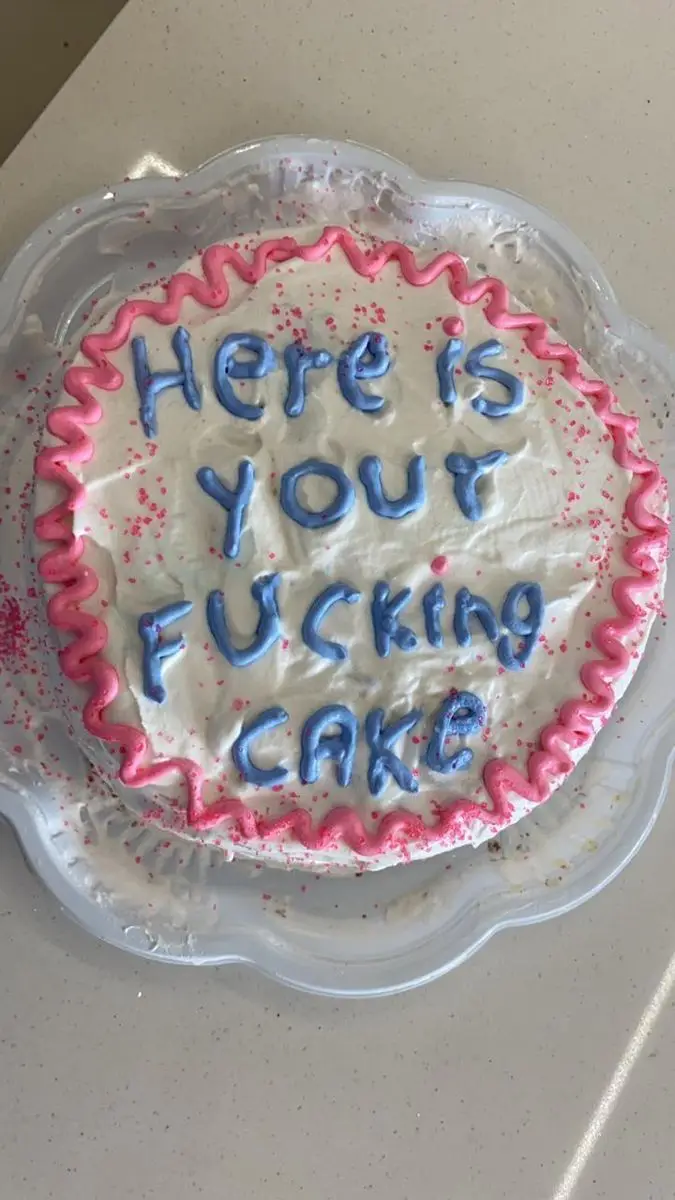 A round white-frosted cake with pink piped icing border and red sprinkles sits on a clear plastic plate. Blue icing letters…