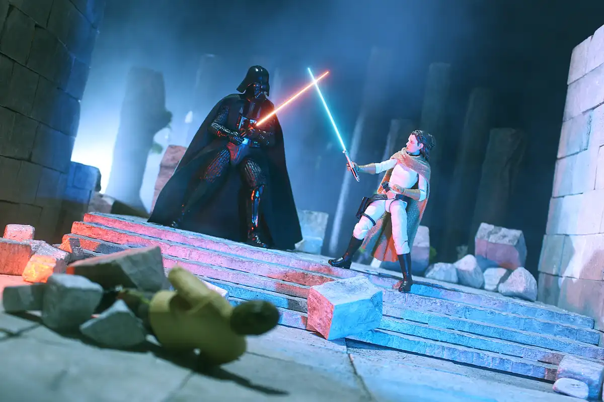 A staged photo of Star Wars action figures: Darth Vader and Rey are locked in a lightsaber duel on stone steps, their blades…