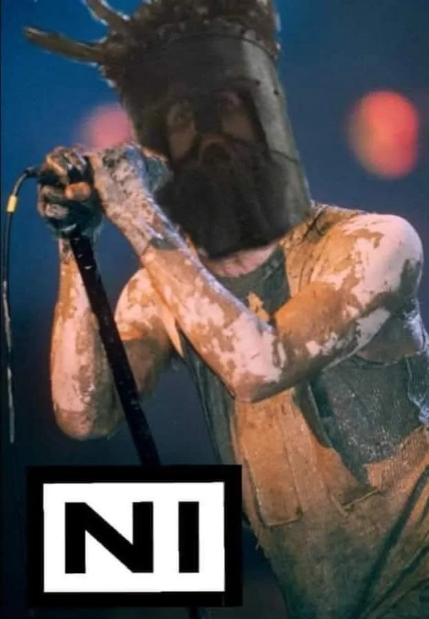 A person covered in mud, wearing a helmet with horns and holding a microphone, with a large white box containing the black…