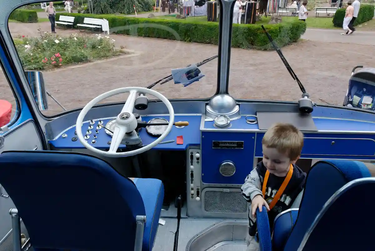 A young child sits in the driver's seat of a vintage blue bus, looking down at the controls with a confused or overwhelmed…