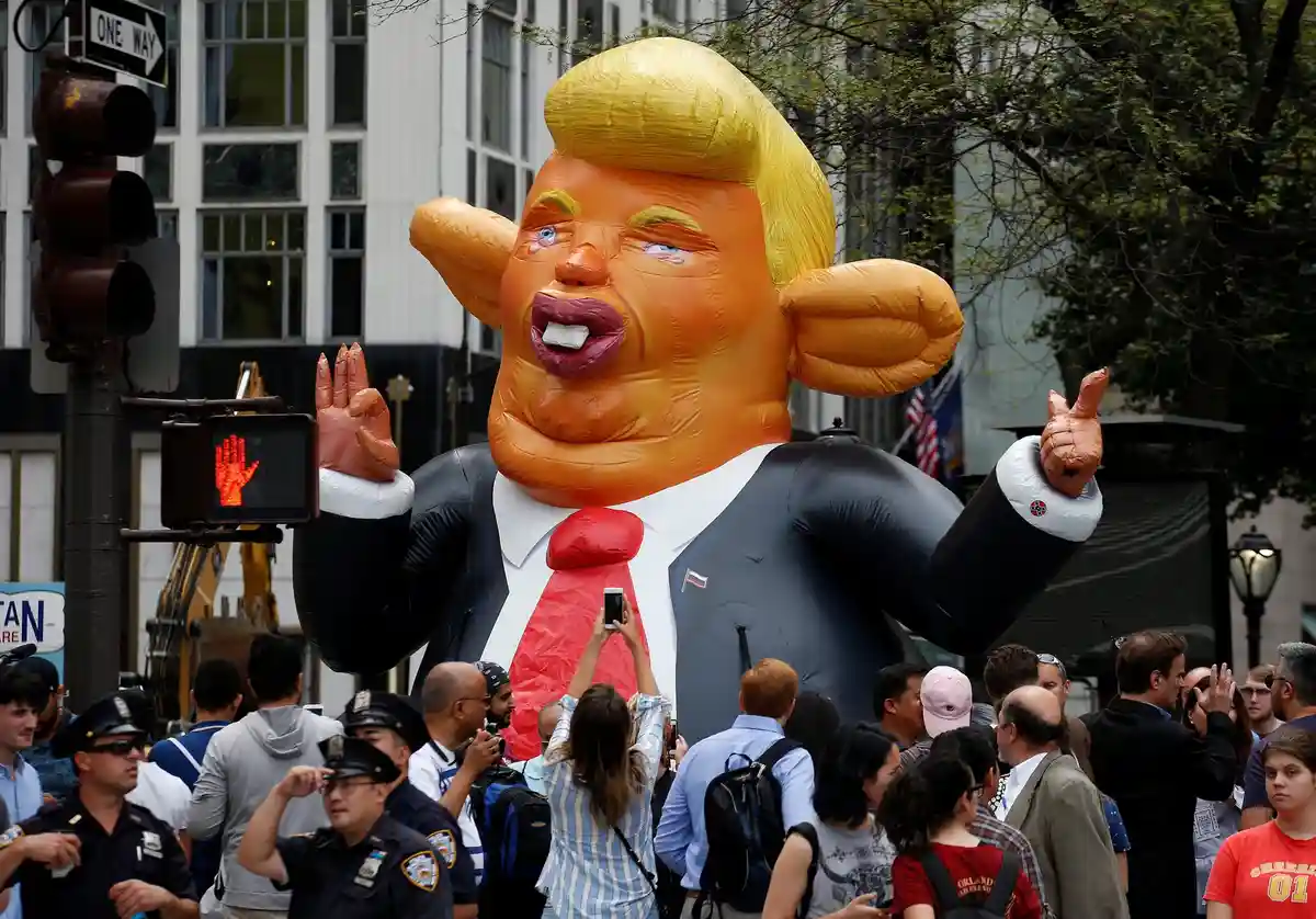 A giant inflatable balloon depicting a man with blonde hair, wearing a suit and red tie, stands in a street with a red hand…