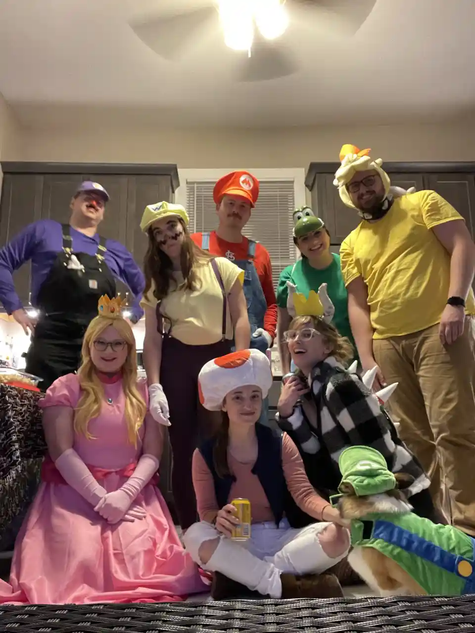 A group of seven people and a dog dressed as Super Mario characters pose for a photo in a kitchen. From left to right: a man…