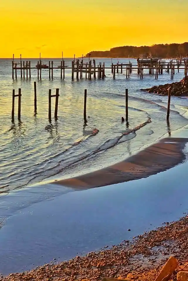 A serene sunset photograph of a beach with wooden pilings extending into calm water, waves gently lapping at the shore…