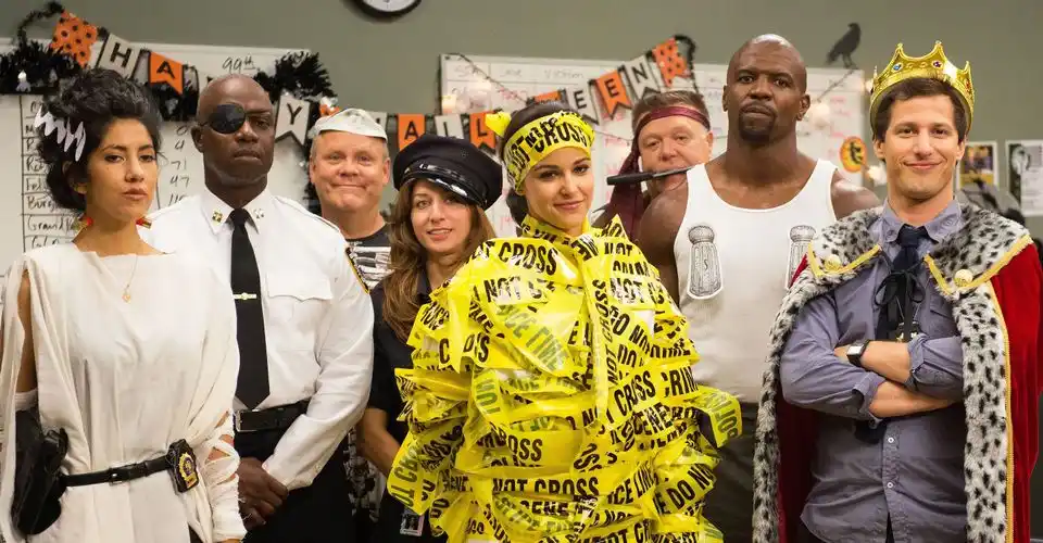 A group of people dressed in Halloween costumes stand in a classroom decorated with orange and black banners. One woman is…
