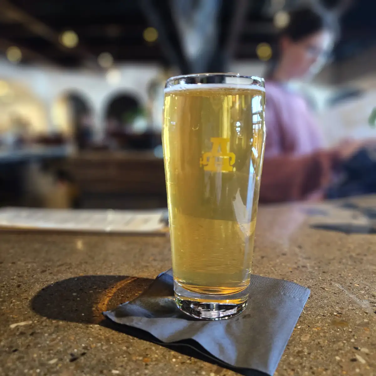 A tall glass of golden beer with a yellow 'A' logo sits on a black napkin on a speckled bar counter. The background is…