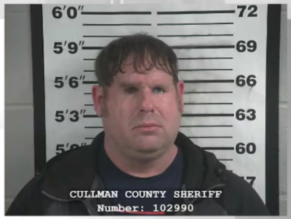 A mugshot of a man with short dark hair and a serious expression, standing in front of a height chart marked in inches and…