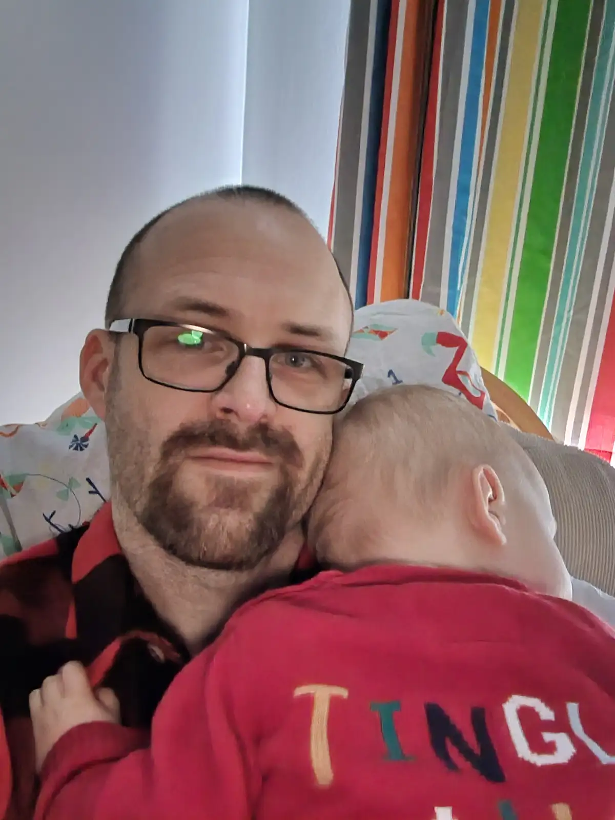 A man with glasses and a beard looks at the camera with a tired, resigned expression while holding a baby who is sleeping on…