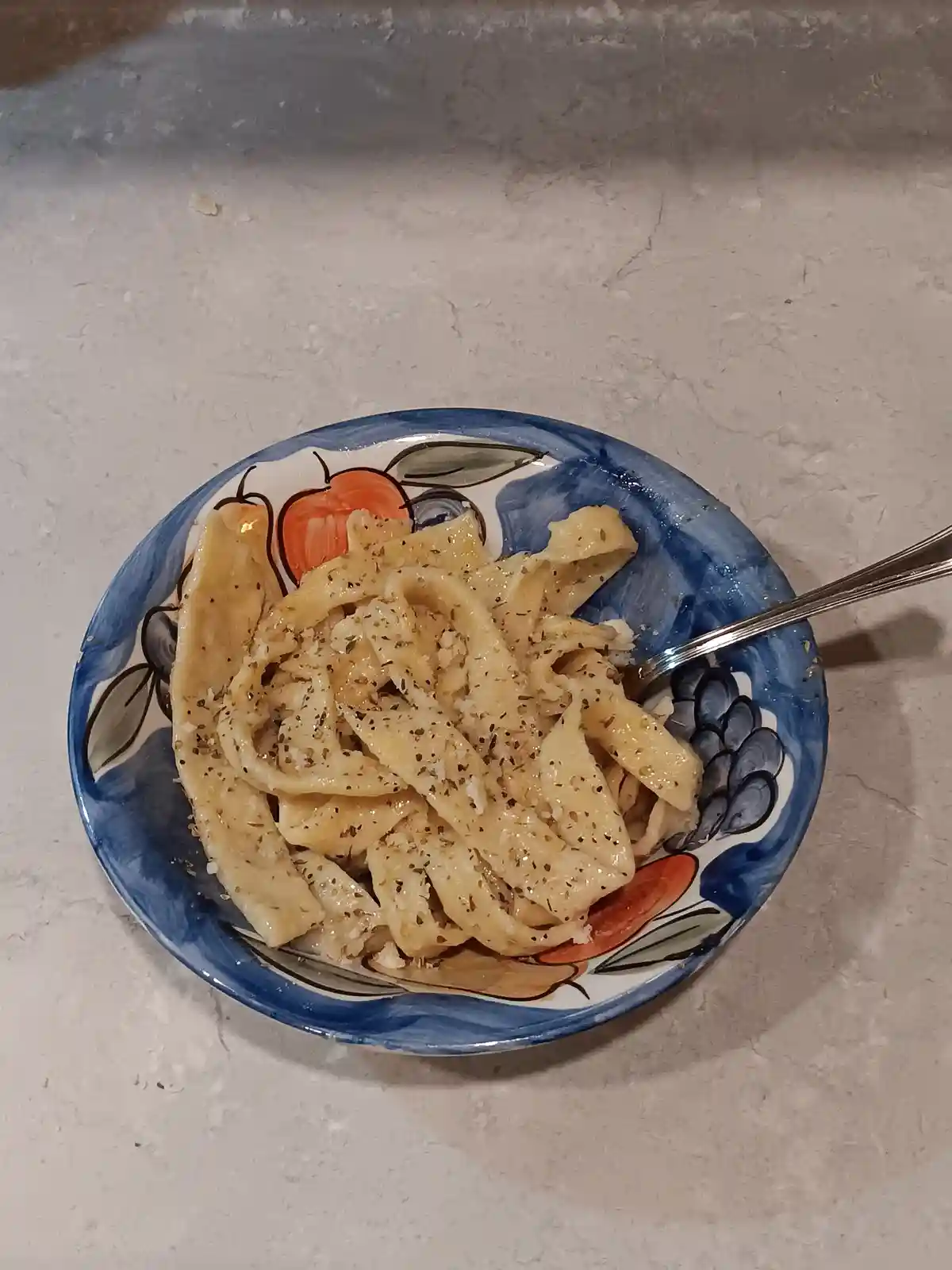 A close-up photo shows a bowl of plain pasta with herbs and pepper, served in a decorative blue and white bowl with fruit…