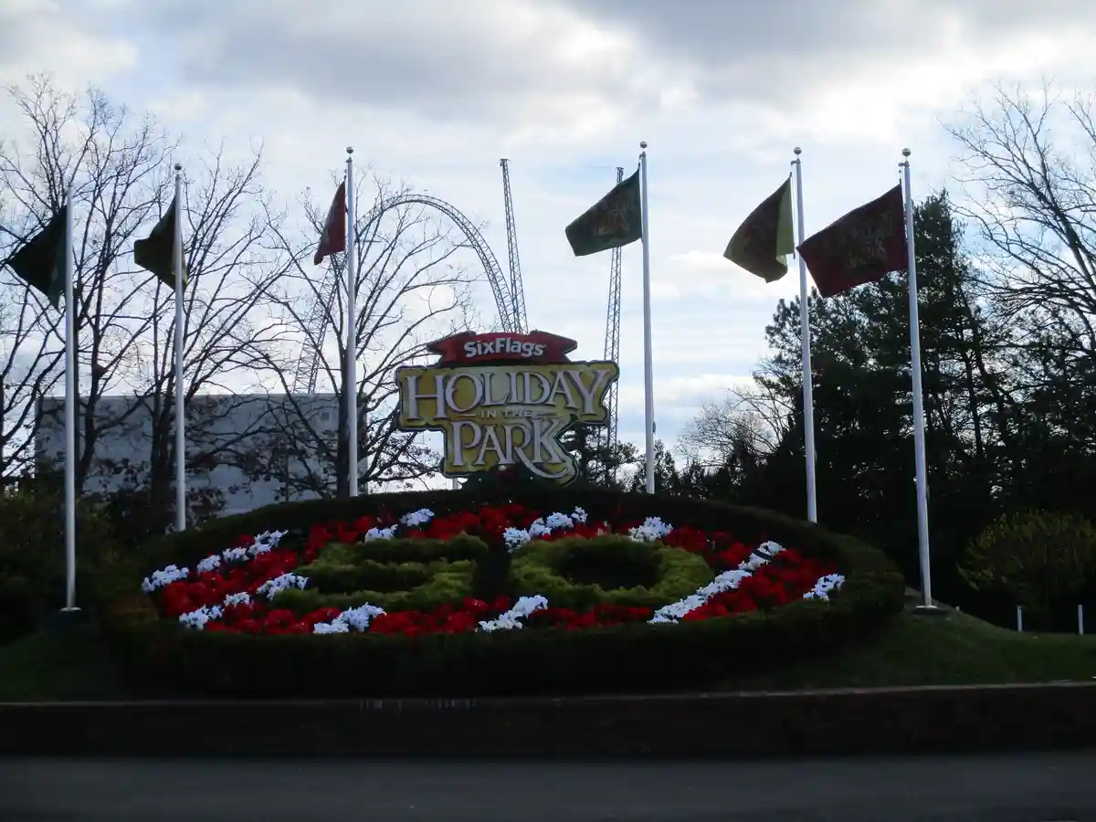 An outdoor photo of a Six Flags 'Holiday in the Park' sign surrounded by red and white flowers arranged in a large, festive…