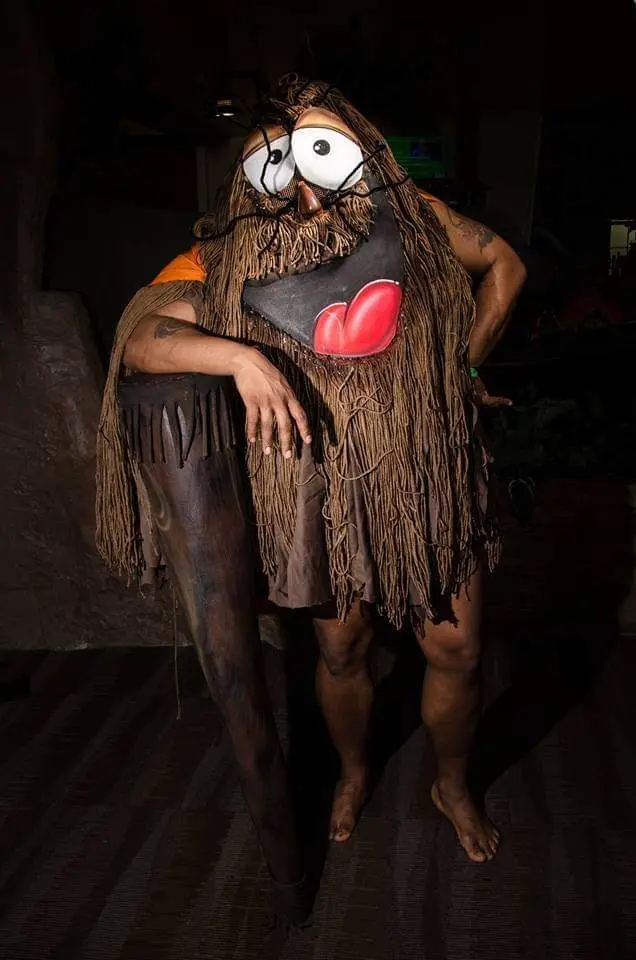 A person in a caveman costume with large cartoon eyes, a brown fringed outfit, and a red heart-shaped mouth mask, poses with…