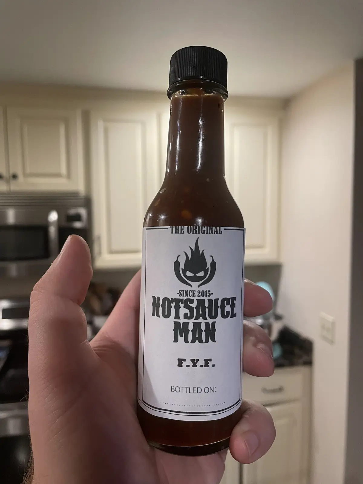 A hand holds a bottle of dark brown hot sauce in a kitchen, the label reads 'THE ORIGINAL -SINCE 2015- HOTSAUCE MAN F.Y.F…