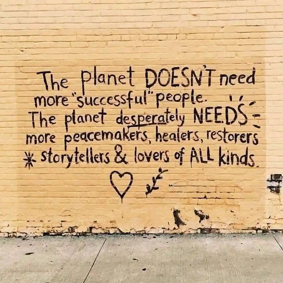 A yellow brick wall has black handwritten text that reads: 'The planet DOESN'T need more "successful" people. The planet…
