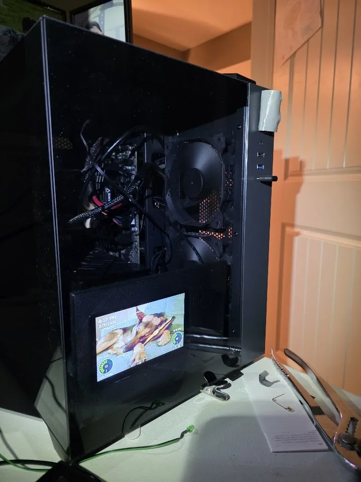 A black computer case with its side panel removed, revealing internal components and a fan. A small screen mounted on the…