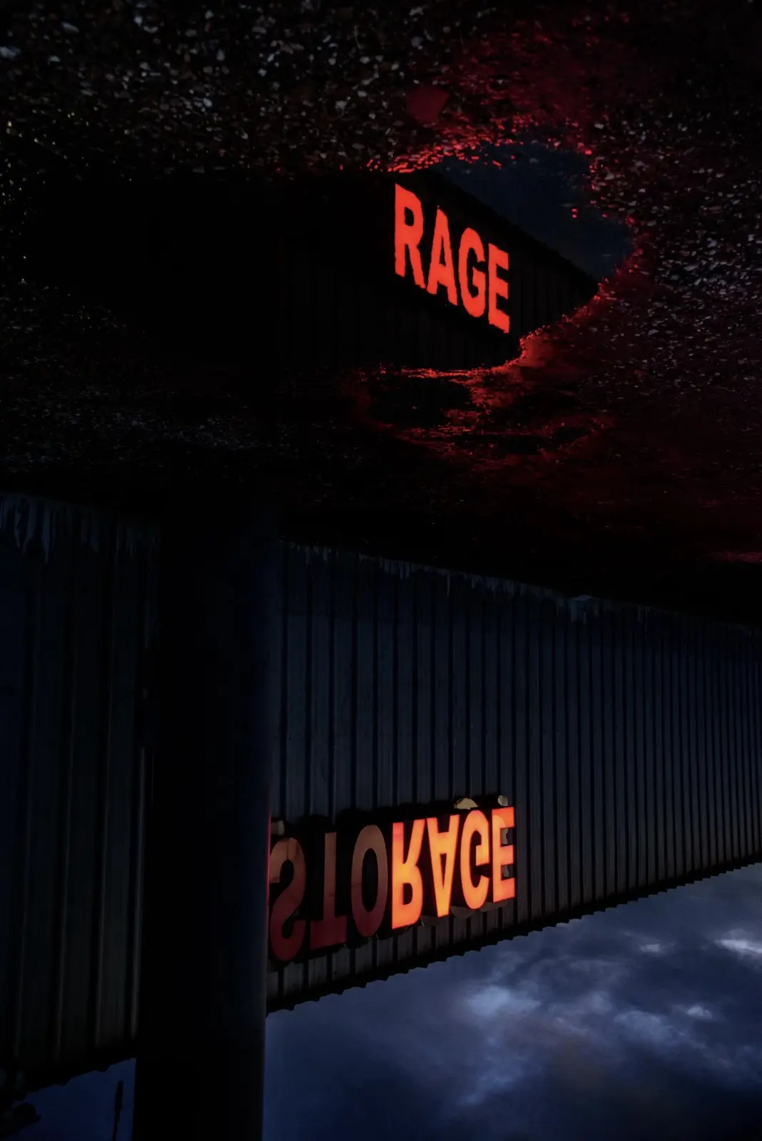 A dark, atmospheric photo shows a corrugated metal building with glowing red neon signs. The top sign reads 'RAGE' and the…