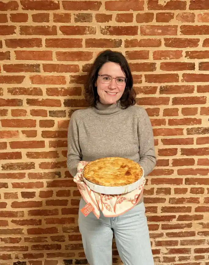 A woman with glasses and a grey turtleneck stands smiling in front of a red brick wall, holding a golden-brown baked pie in…