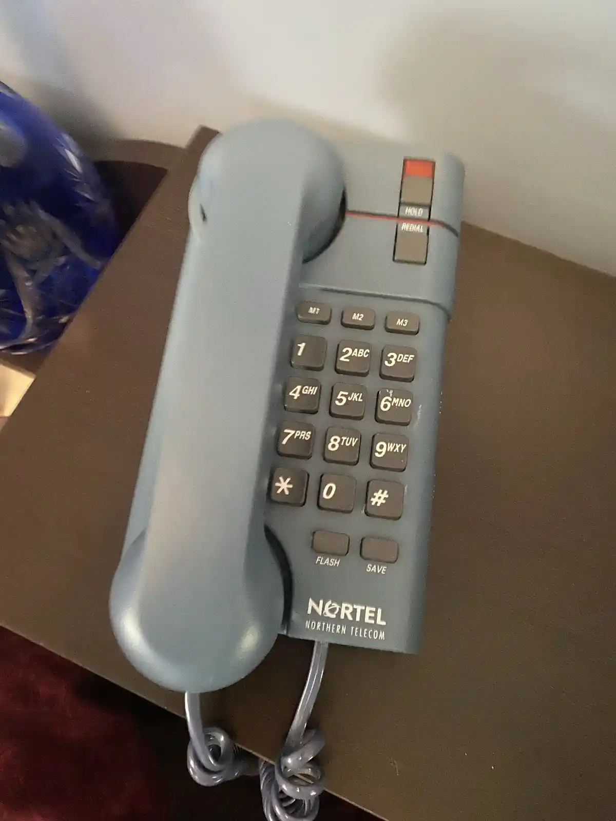 A close-up photo of a grey, corded Nortel Northern Telecom landline phone sitting on a brown surface, with a blue plastic…