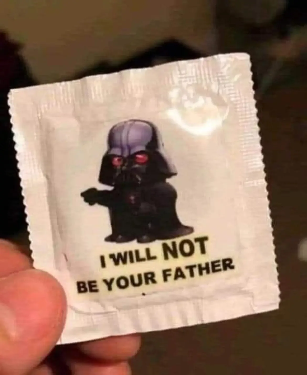 A close-up photo shows a hand holding a white condom wrapper. The wrapper features a small, chibi-style Darth Vader figure…
