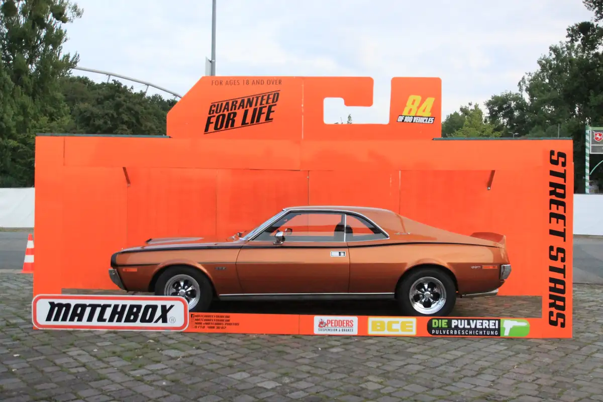 A real orange muscle car is displayed outdoors inside a giant orange cardboard box resembling a Matchbox toy car package…