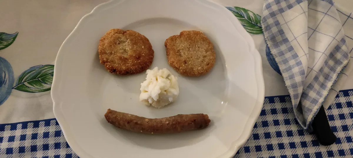 A white plate holds two round fried items, a dollop of mashed potatoes, and a sausage arranged to resemble a smiling face…