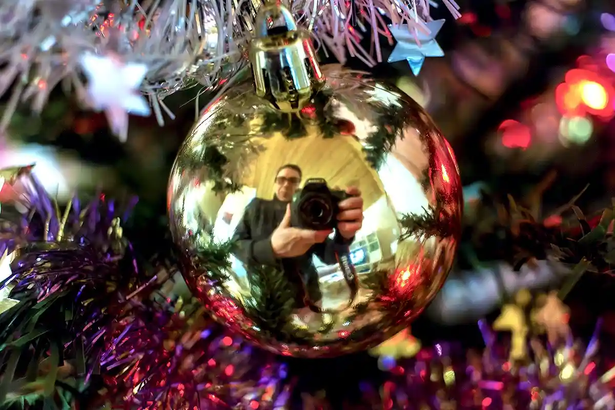 A shiny gold Christmas ornament hangs on a decorated tree, reflecting the image of a person holding a camera to take a…