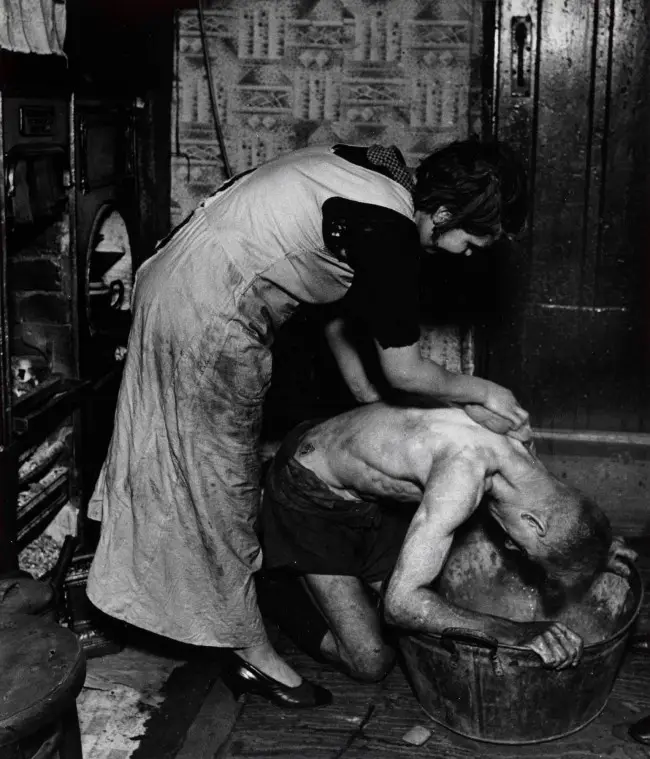 A black-and-white photo shows a woman in an apron scrubbing a shirtless man's back while he kneels over a large metal tub…