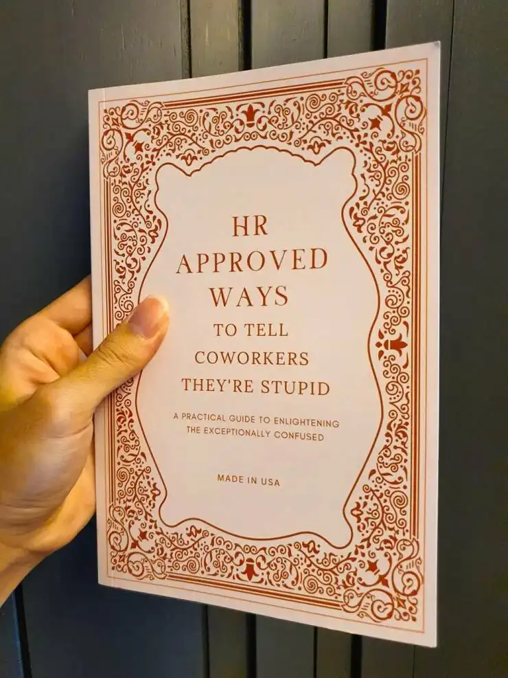 A hand holds a book with an ornate, vintage-style red border. The cover reads 'HR APPROVED WAYS TO TELL COWORKERS THEY'RE…