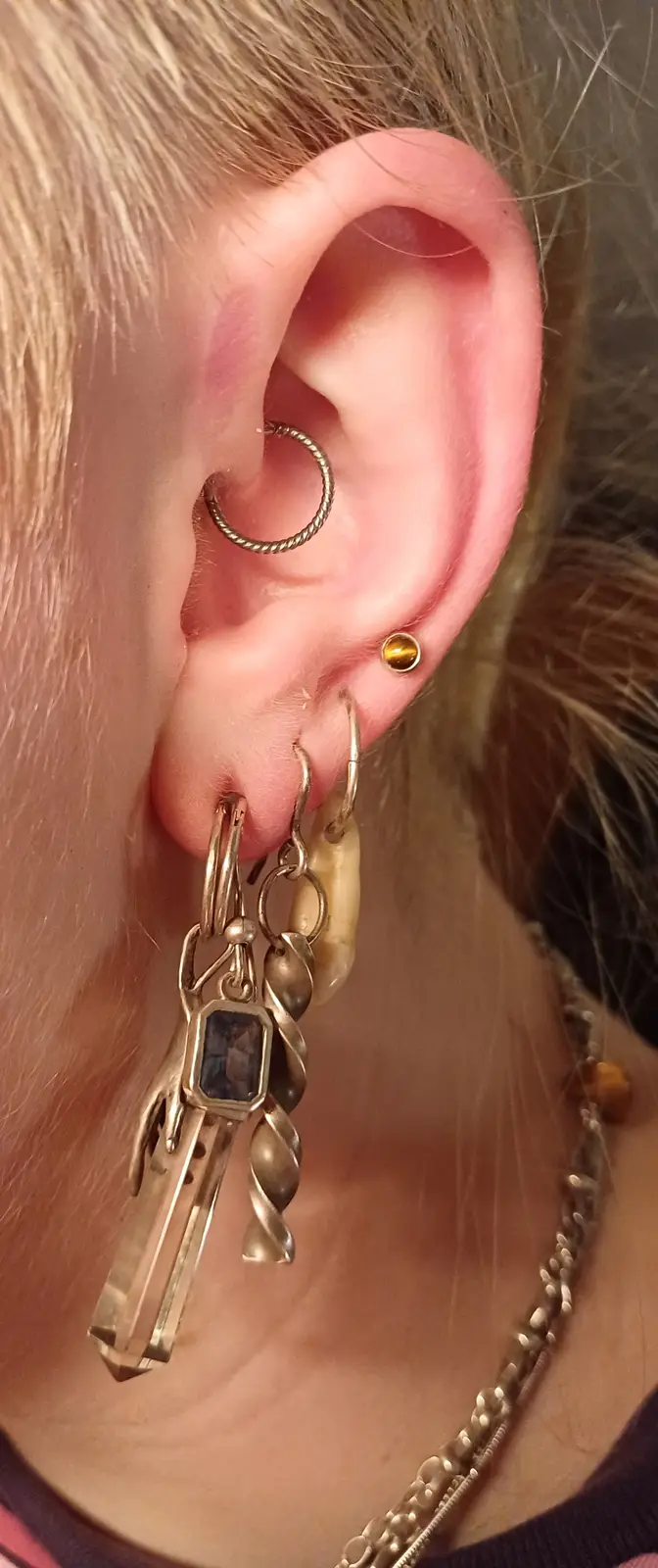 A close-up photo of a person's ear with multiple piercings, including a snake chain hoop, a small gold ball stud, and a…