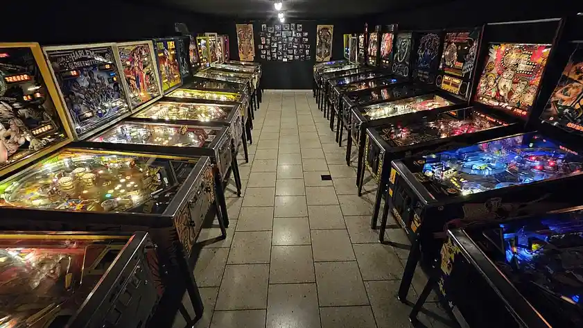 A long, dimly lit hallway lined with dozens of brightly lit pinball machines on both sides, their colorful backglass artwork…
