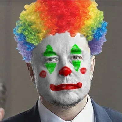 A man in a suit and white face paint with green triangles, red nose, and red lips wears a rainbow-colored curly wig. The…