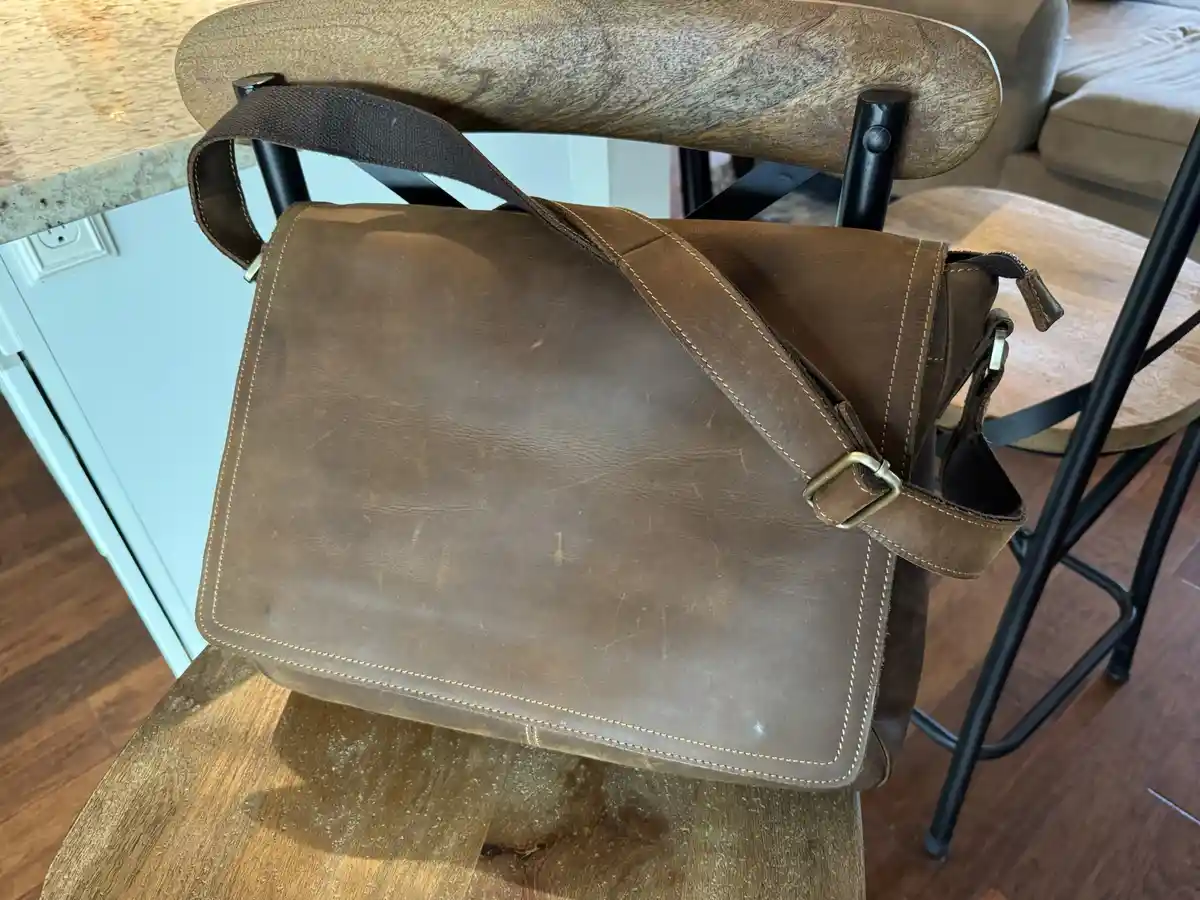 A well-worn brown leather messenger bag with visible scuffs and light-colored stitching is draped over the back of a wooden…