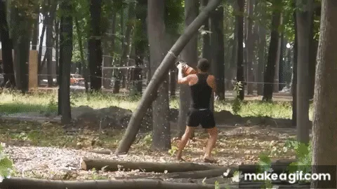A man in a black tank top and shorts is shown in a sequence of frames performing pull-ups on a wooden bar in a wooded area…