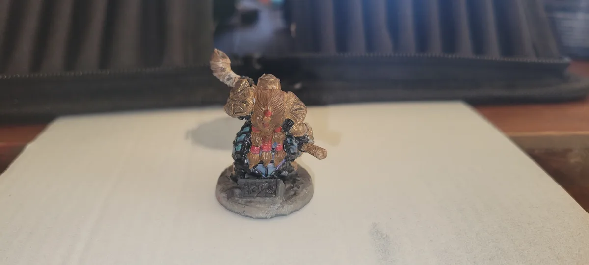A hand-painted fantasy miniature figure, likely a dwarf or warrior, stands on a grey circular base on a white surface. The…