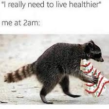A raccoon is shown walking on a sandy surface while carrying a large, red-and-white striped bag of chips in its paws. The…